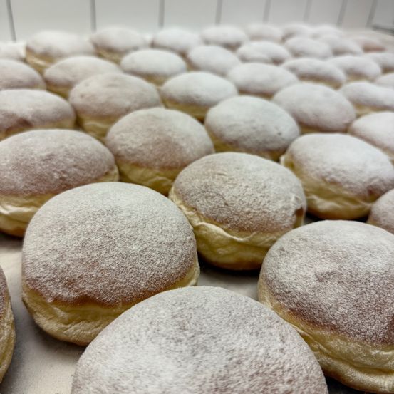 Ein Nahaufnahme einer mit zahlreichen mit Puderzucker bestreuten Donuts gefüllten Schale. Die Donuts haben eine hellbraune Farbe und sind in Reihen angeordnet.