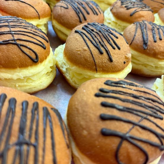 Eine Nahaufnahme mehrerer Gebäckstücke, jedes mit einer Spirale aus Schokoladenglasur gekrönt. Die Gebäckstücke haben eine goldbraune Kruste und eine cremige Füllung, möglicherweise mit Pudding.