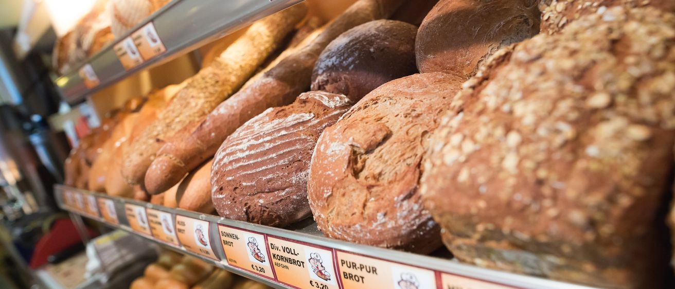 Ein Bäckereiregal zeigt verschiedene Brotsorten, darunter Roggenbrot, Vollkornbrot und Sauerteigbrot, mit Preisschildern und einem Preis von 3,20 Euro.