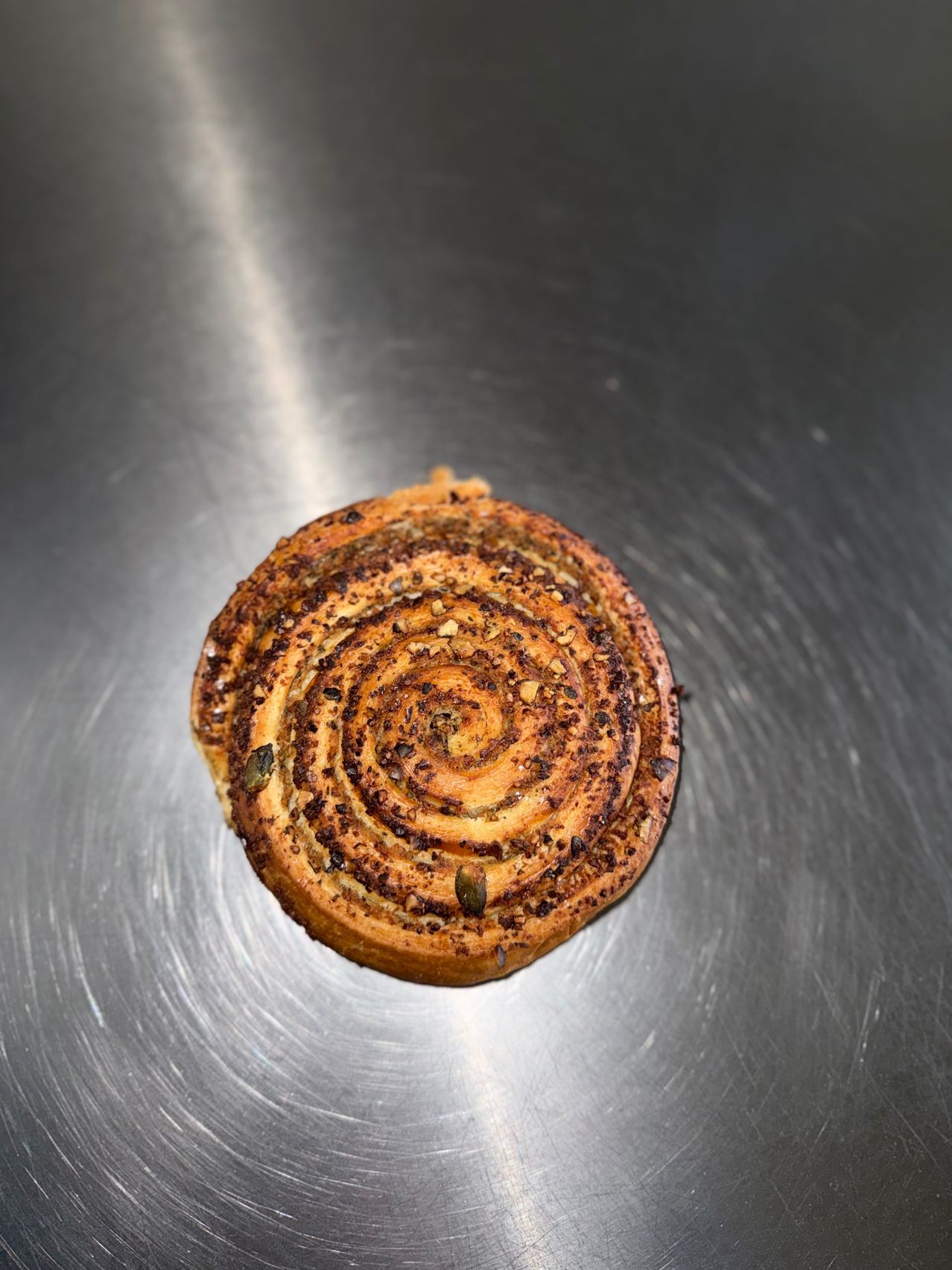 Ein Spiralgebäck mit goldbrauner Kruste und sichtbaren Samen, auf einer metallischen Oberfläche.
