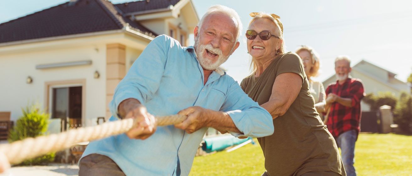 Ein Paar, das vor einem Haus auf einem Rasen einen fröhlichen Tauziehen veranstaltet. Der ältere Mann und die Frau lächeln und halten ein Seil, während andere im Hintergrund zusehen.