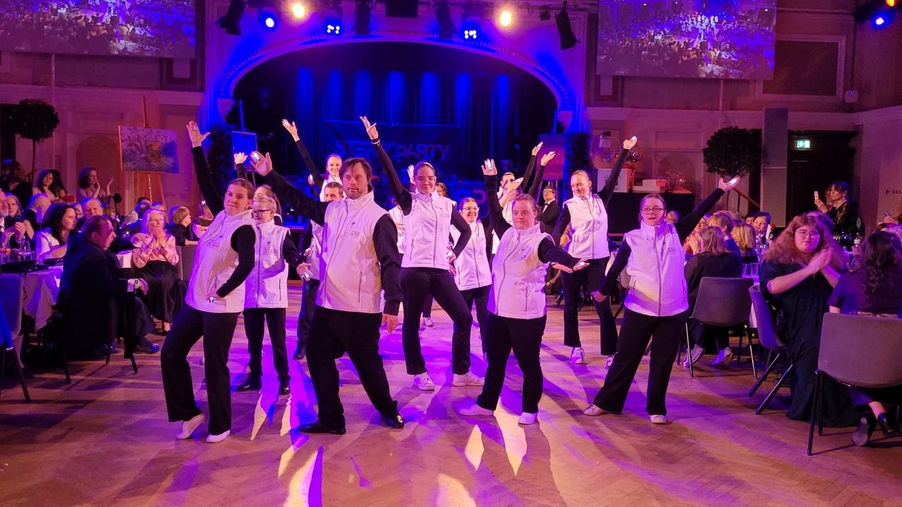 Eine Gruppe von Menschen, die in Weiß und Schwarz gekleidet sind, führt einen Tanz auf einer Bühne mit lila Beleuchtung auf. Einige von ihnen tragen Brillen und einige Armbänder. Dahinter sind verschwommene Bilder von Zuschauern zu sehen.