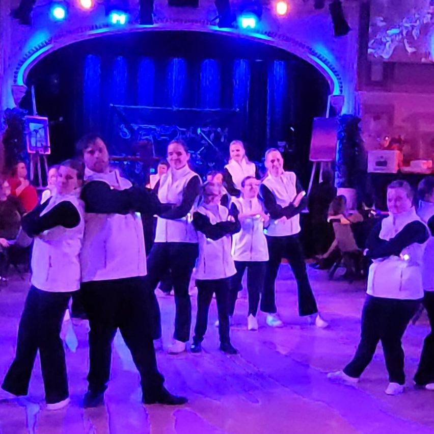 Eine Gruppe von Menschen in weißen und schwarzen Outfits tritt auf einer Bühne mit lila Lichtern auf. Sie stehen in einer Reihe, und einige tanzen. Im Hintergrund sitzen andere Leute und schauen zu.