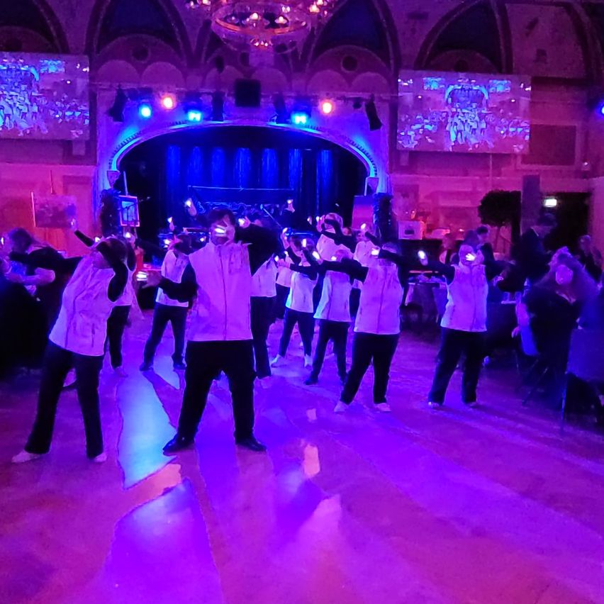 Eine Gruppe von Menschen in gleichen weißen Outfits führt einen synchronisierten Tanz unter lila Lichtern in einem Ballsaal auf.