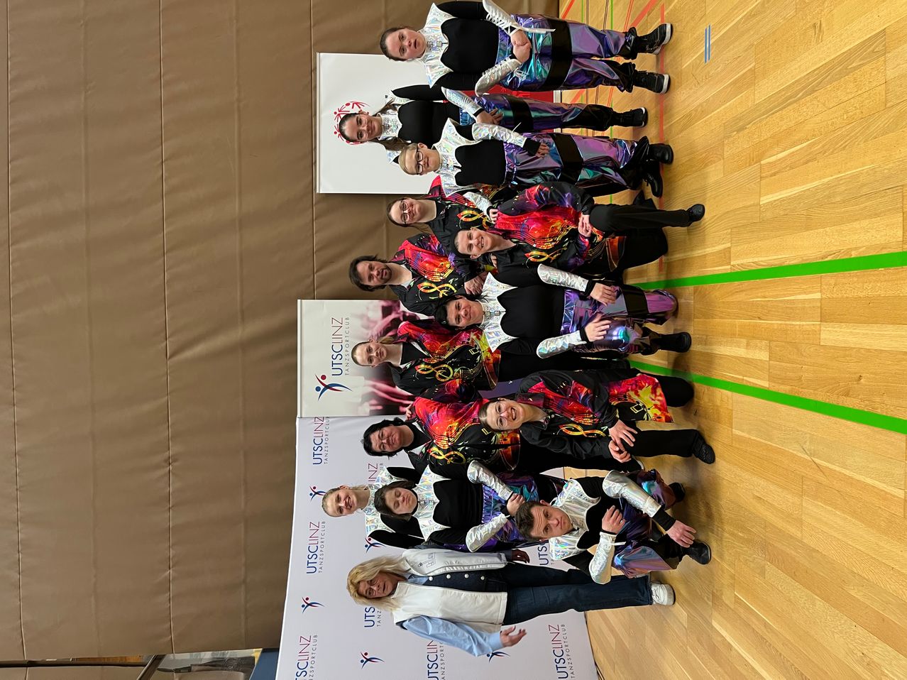Eine Gruppe von Menschen in farbenfrohen, leuchtenden Kostümen posiert für ein Foto in einer Turnhalle. Ein Banner mit UTSCLNZ und einem Logo ist im Hintergrund sichtbar.