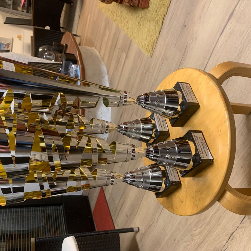 Eine Ausstellung von Silber- und Goldtrophäen auf einem runden Holztisch mit gelber Basis und schwarzen Akzenten. Der Raum hat Holzboden.