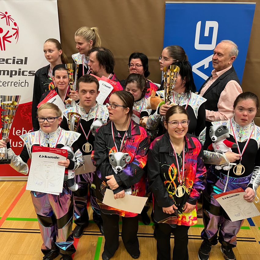 Eine Gruppe von Menschen steht in einem Fitnessstudio zusammen, alle in farbenfrohen Kostümen. Sie halten Urkunden und Medaillen und einige tragen eine Brille. Eine Person vorne hält ein Kuscheltier. Hinter ihnen steht ein blauer Banner mit Text darauf.
