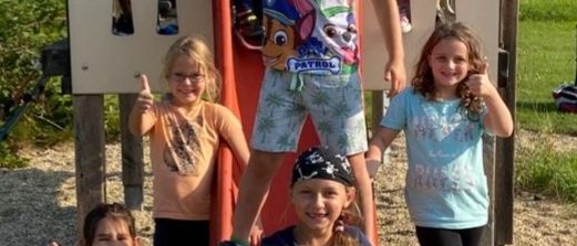 Eine Gruppe von Kindern spielt auf einem Spielplatz. Einige sitzen auf einer Rutsche, während andere darum herumstehen.