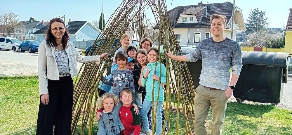 Eine Gruppe von Menschen, einschließlich Erwachsener und Kindern, posiert für ein Foto vor einer großen, handgefertigten Struktur aus Stöcken auf einer Rasenfläche.