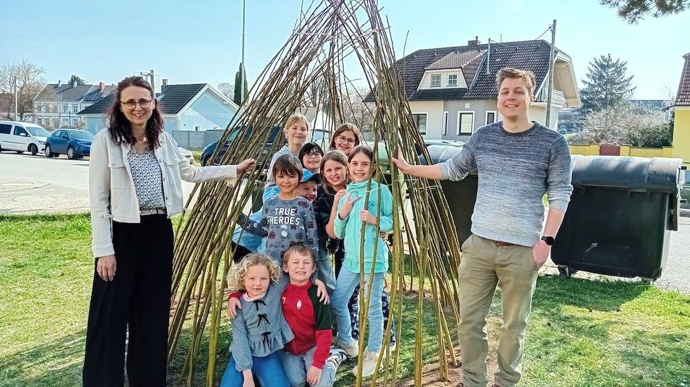 Eine Gruppe von Menschen, einschließlich Erwachsener und Kindern, posiert für ein Foto vor einer großen, handgefertigten Struktur aus Stöcken auf einer Rasenfläche.