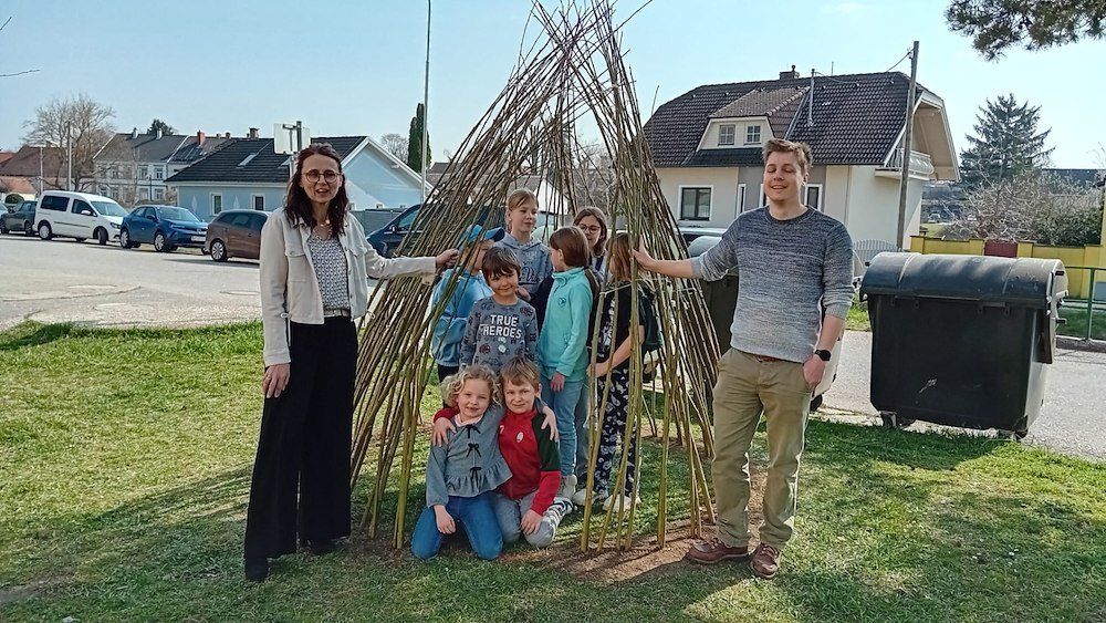 Eine Gruppe von Menschen, einschließlich Erwachsener und Kinder, steht vor einer handgefertigten Struktur aus Stöcken in einem Park.