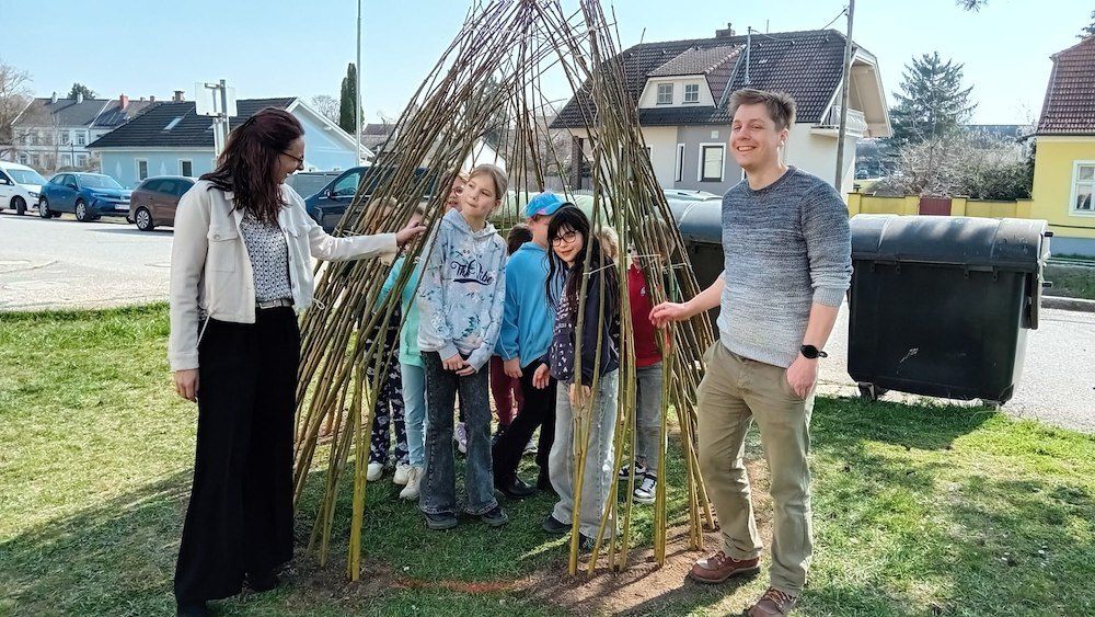 Eine Gruppe von Kindern und zwei Erwachsene stehen um eine Struktur aus Holzstäben auf einem Rasenfeld.