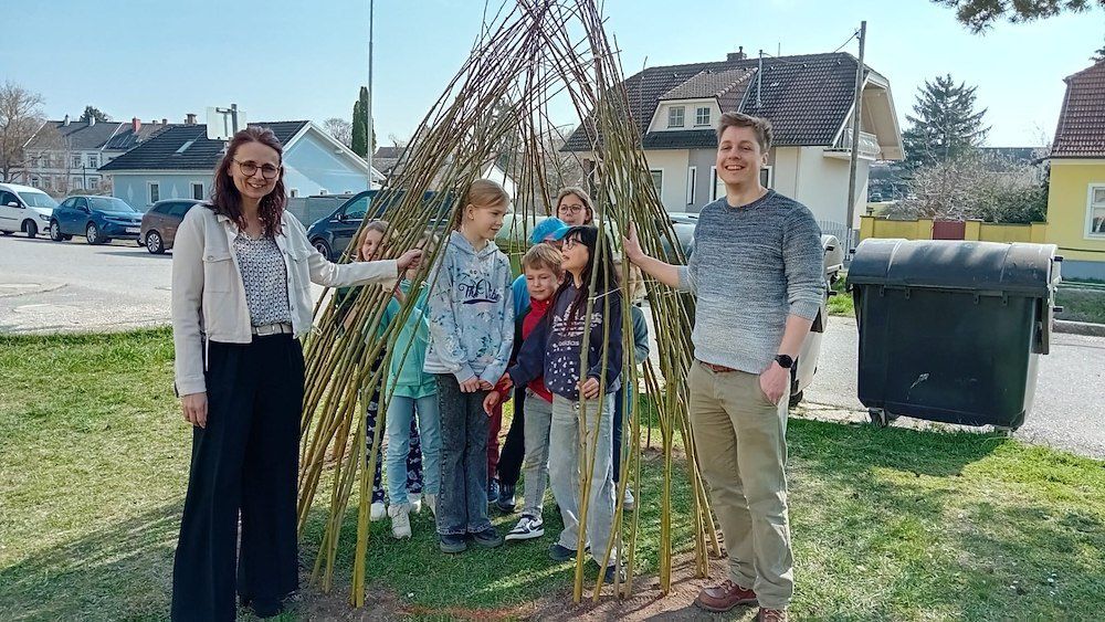 Eine Gruppe von Kindern und zwei Erwachsene stehen vor einer großen Bambusstruktur in einem Park.
