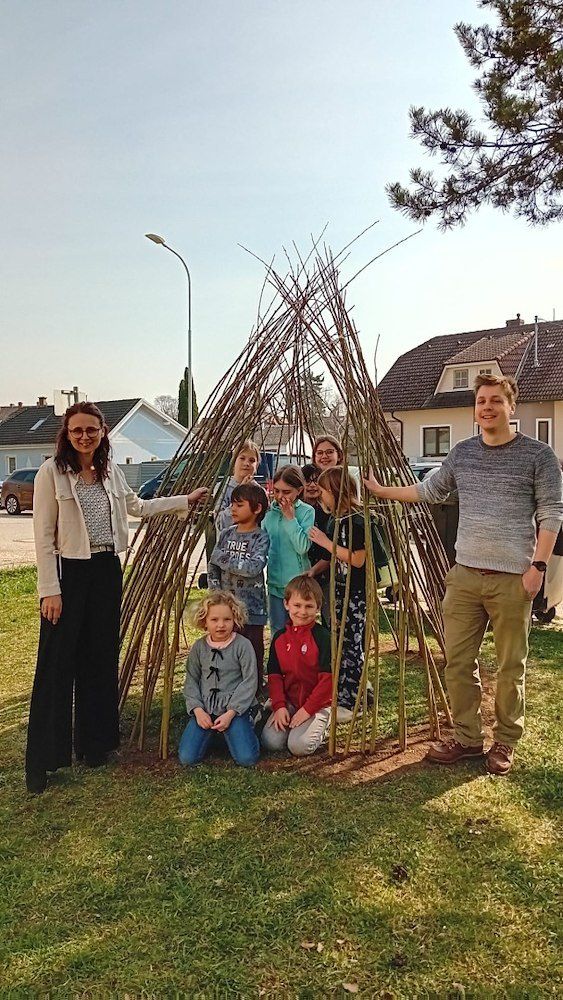 Eine Gruppe von Menschen, einschließlich Erwachsener und Kindern, steht um eine Struktur aus Stöcken herum. Sie posieren für ein Foto in einem Grasbereich.