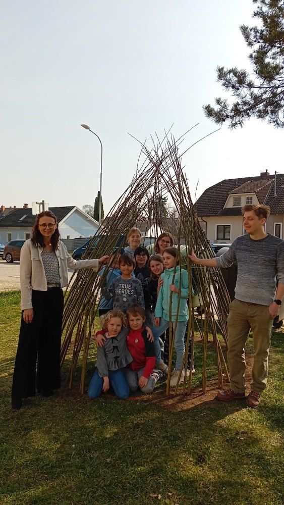 Eine Gruppe von Menschen, einschließlich Erwachsener und Kindern, posiert für ein Foto vor einer handgefertigten Struktur aus Stöcken in einem Grasbereich.
