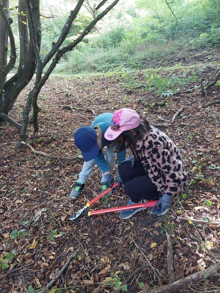 Zwei Kinder sind in einem Wald, eines benutzt eine rote Gartenschere, um einen Ast zu schneiden, während das andere auf den Boden schaut.