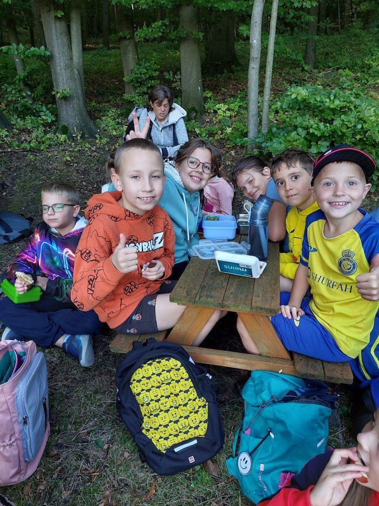Eine Gruppe von Kindern sitzt um einen Picknicktisch in einem Wald. Sie lächeln und scheinen ihr Mittagessen zu genießen.