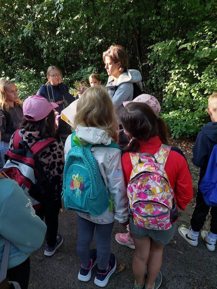 Eine Gruppe von Kindern und Erwachsenen steht in einem Wald, mit einer Frau in der Mitte, die ein Blatt hält.