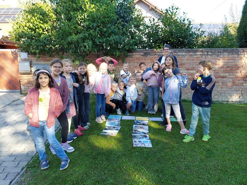 Eine Gruppe von Kindern und Erwachsenen steht auf einem Rasen und posiert für ein Foto. Einige der Kinder tragen eine Brille und einige tragen Turnschuhe. Sie stehen vor einer Ziegelmauer und hinter ihnen steht ein Baum mit grünen Blättern.
