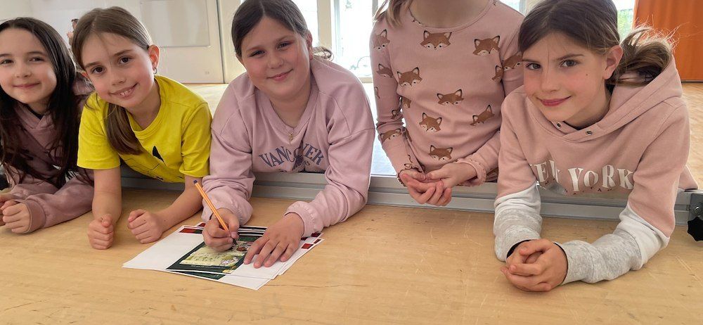 Fünf junge Mädchen sitzen an einem Tisch, lächeln und posieren für ein Foto. Ein Mädchen hält einen Stift und scheint auf einem Stück Papier zu zeichnen.