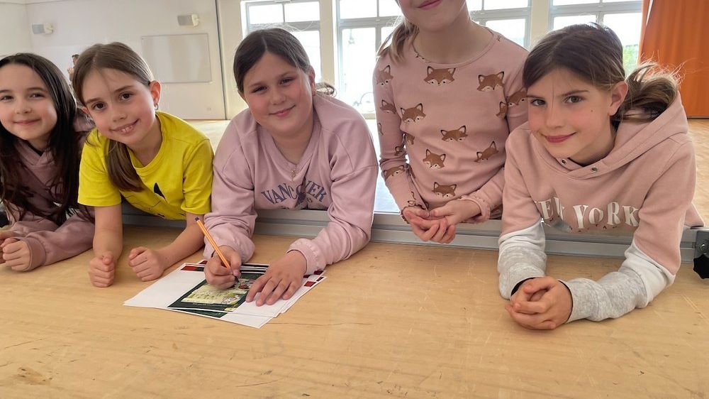 Fünf junge Mädchen sitzen an einem Tisch, lächeln und posieren für ein Foto. Ein Mädchen hält einen Stift und scheint auf einem Stück Papier zu zeichnen.