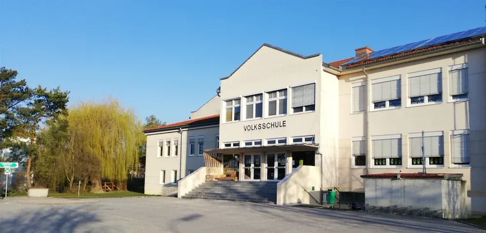 Ein weißes Gebäude mit einem roten Dach und Solarpanelen auf der Oberseite. Das Gebäude hat viele Fenster und ein Schild, das 'Volksschule' besagt.