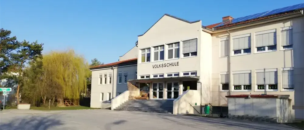Die Vorderseite eines weißen Gebäudes mit der Aufschrift Volksschule über dem Eingang. Es hat Glasfenster und Türen und ist von Bäumen und Gras umgeben.