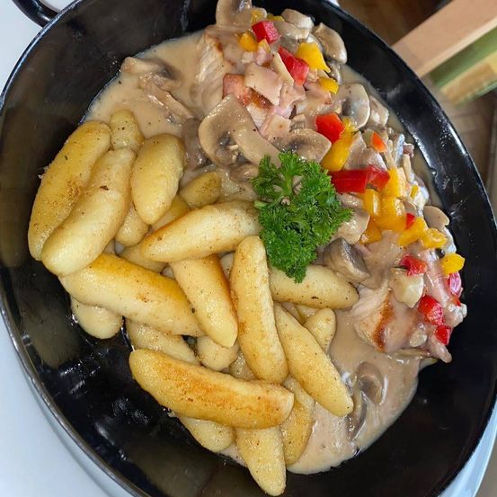 Eine schwarze Pfanne gefüllt mit einem cremigen Champignon- und Paprikagericht, begleitet von goldenen Kartoffeln und einem Petersilienzweig.