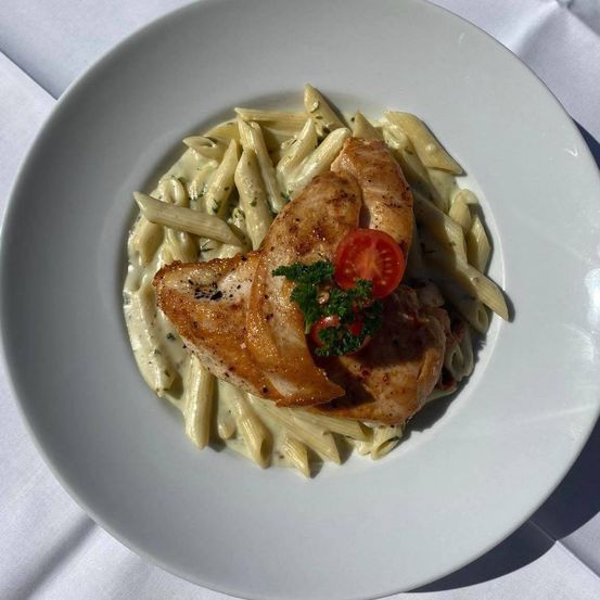Ein weißer Teller mit einem Gericht aus Penne-Pasta mit cremiger Sauce, belegt mit zwei Stücken gegrilltem Hähnchen und einer Scheibe Tomate.