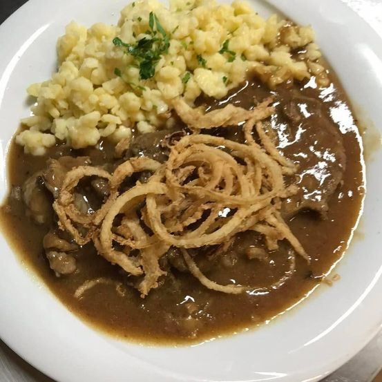 Ein weißer Teller mit einem herzhaften Gericht aus Rindergulasch, überzogen mit knusprigen Zwiebelringen und serviert mit cremigen Kartoffelpüree.