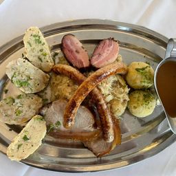 Ein silberner Teller mit einer Auswahl an deutschen Gerichten, darunter Würste, Sauerkraut und Knödel, begleitet von einem Soßenkessel.
