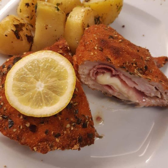 Ein Teller mit zwei panierten Hähnchenschnitzeln, eines davon mit einer Zitronenscheibe garniert, und einer Beilage aus gebratenen Kartoffeln.