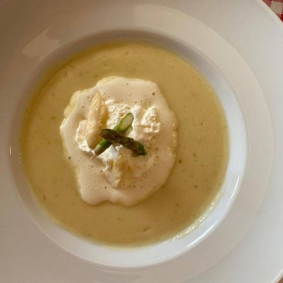 Eine Schüssel mit cremiger Suppe, gekrönt von weißer Sahne und Spargelstücken. Die Suppe hat eine hellgelbe Farbe.