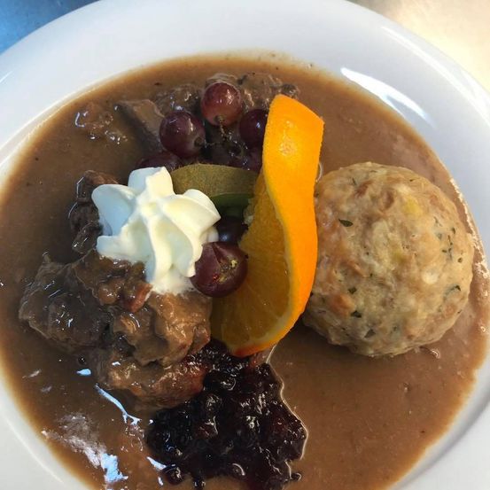 Ein weißer Teller mit einem Gericht aus geschmortem Fleisch in Soße, garniert mit einem Klecks saure Sahne, einer Scheibe Orange und einer Fleischknödel, begleitet von einer Beilage aus Cranberries.