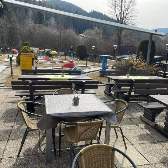 Eine Außengastronomie mit Tischen und Stühlen, einem Minigolfplatz im Hintergrund und einer malerischen Aussicht auf Berge und Bäume.