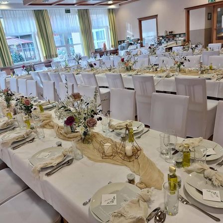 Ein langer Banketttisch für eine große Veranstaltung, mit weißen Stühlen, weißer Tischdecke und Blumengestecke. Der Tisch ist mit Tellern, Gläsern und Besteck geschmückt, und es gibt Kerzen und Flaschen auf dem Tisch. Der Raum hat große Fenster mit Vorhängen und eine Holztür.