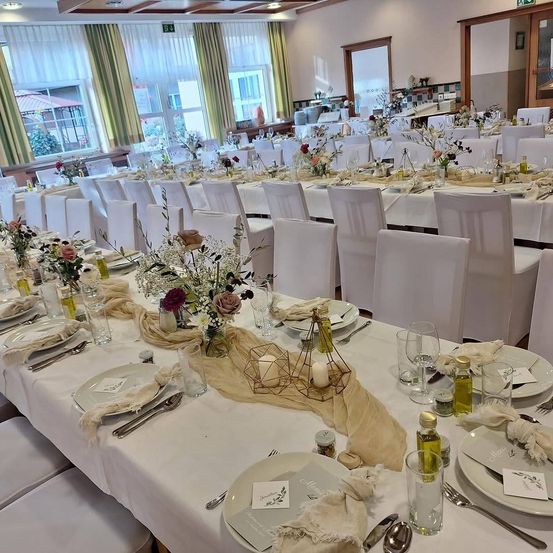 Ein langer Banketttisch für eine große Veranstaltung, mit weißen Stühlen, weißer Tischdecke und Blumengestecke. Der Tisch ist mit Tellern, Gläsern und Besteck geschmückt, und es gibt Kerzen und Flaschen auf dem Tisch. Der Raum hat große Fenster mit Vorhängen und eine Holztür.