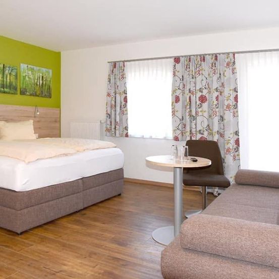Ein Schlafzimmer mit einem Doppelbett, einem kleinen Tisch mit einem Handy, einem Stuhl und einer Couch in der Nähe eines Fensters mit Blumenvorhängen. Die Wände sind grün gestrichen, und es gibt drei Gemälde an der Wand.