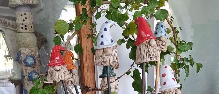 Eine Ausstellung von Gartenzwergen und anderen Tonfiguren mit einer Pflanze in einem Korb an einem Schaufenster. Verschiedene Tonobjekte sind im Fenster reflektiert.