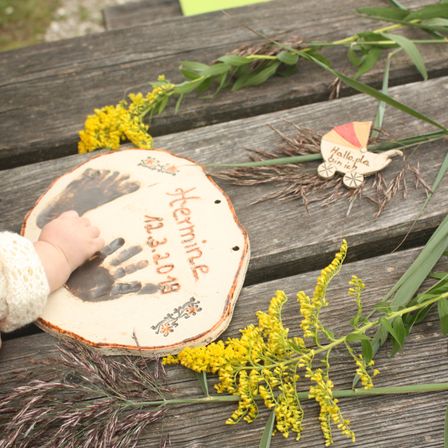 Der Hand- und Fußabdruck eines Babys auf einer Holzscheibe, umgeben von gelben Blumen und Grünpflanzen, auf einem Holztisch.