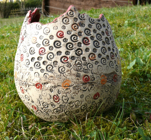 Ein großes Keramikobjekt mit rissiger Oberfläche und farbenfrohen Spiralmustern liegt auf einem Grasfeld. Das Objekt ist weiß mit roten, schwarzen und orangefarbenen Spiralen.