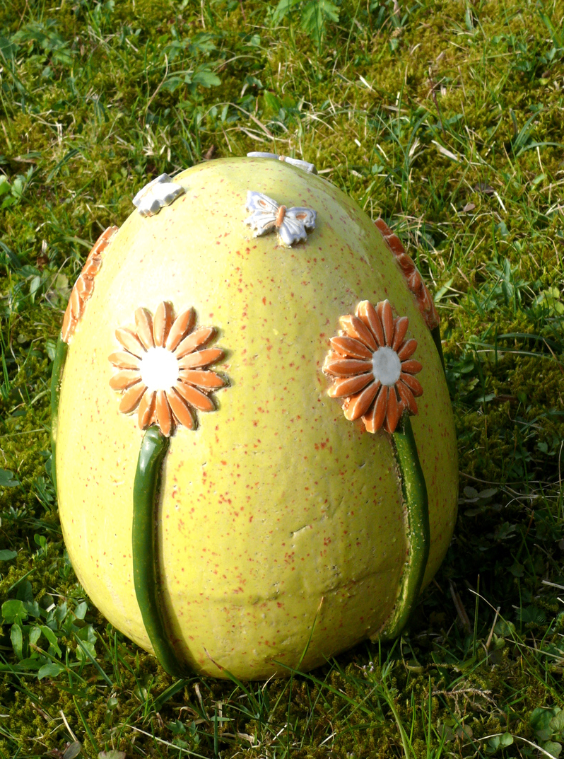 Ein großes gelbes Ei mit roten Flecken, verziert mit drei orangefarbenen Blumen und zwei Schmetterlingen auf der Oberseite. Es steht auf grünem Gras.