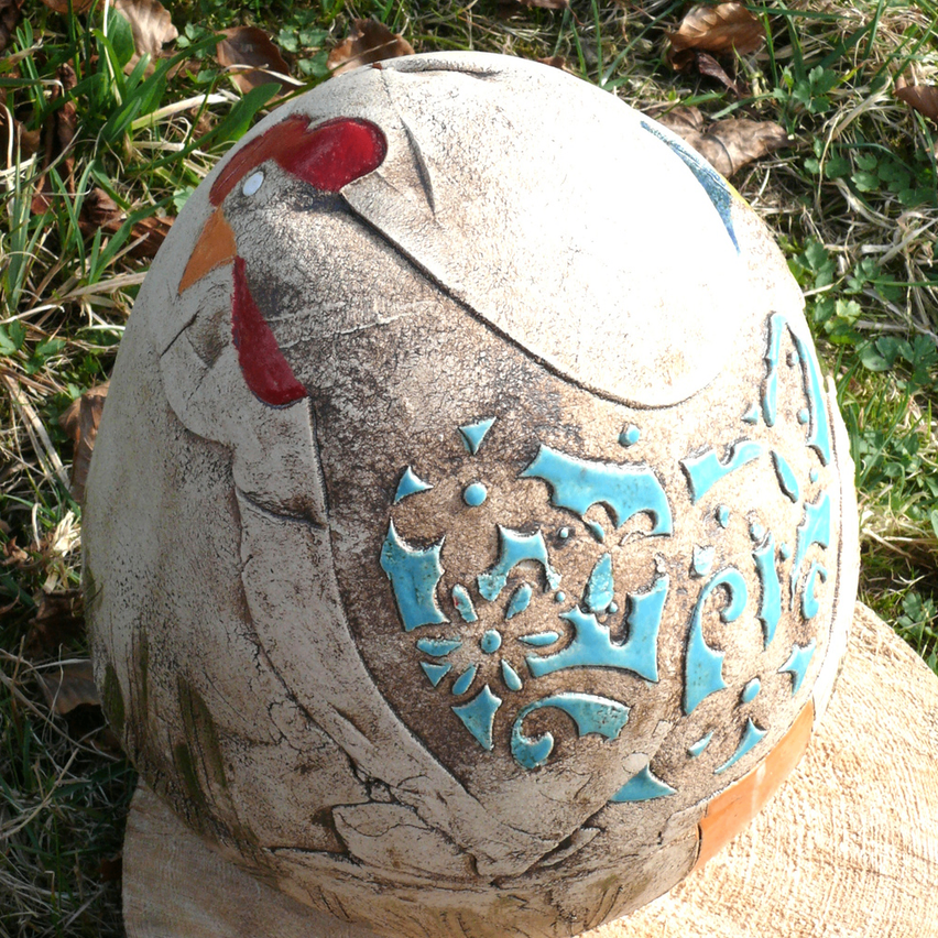 Eine weiße und blaue Keramik-Hahn-Skulptur steht auf einer Holzoberfläche, draußen auf einer Grasfläche mit trockenen Blättern.