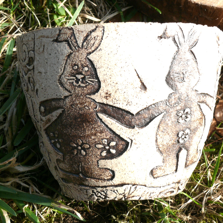 Ein Keramiktopf mit zwei Händchen haltenden Kaninchen, eines mit Blumen, steht auf Gras. Der Topf ist hellbraun mit dunkleren braunen Details.