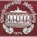 Ein roter Hintergrund zeigt ein klassisches architektonisches Emblem. Die Struktur ist symmetrisch mit mehreren Säulen und einer zentralen Kuppel, umgeben von Blättern. Die Worte 'Wandelceunde Helfensburg' sind in Weiß oben geschrieben, und das Jahr '1975' ist darunter eingraviert.