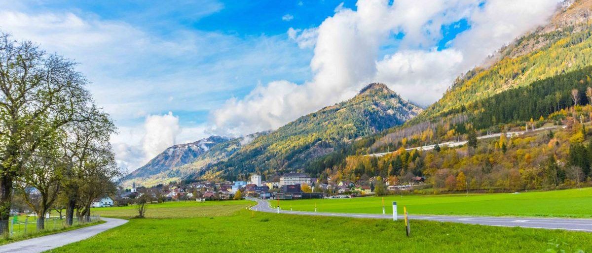 Eine ländliche Landschaft mit einem Bergrücken im Hintergrund. Eine Straße schlängelt sich durch ein grünes Feld. Mehrere Gebäude sind verstreut.