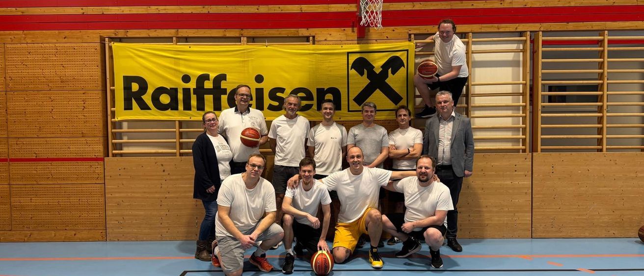 Eine Gruppe von Menschen posiert für ein Foto in einer Turnhalle mit einem Basketballplatz. Ein Banner mit der Aufschrift 'Raiffeisen' hängt über ihnen.