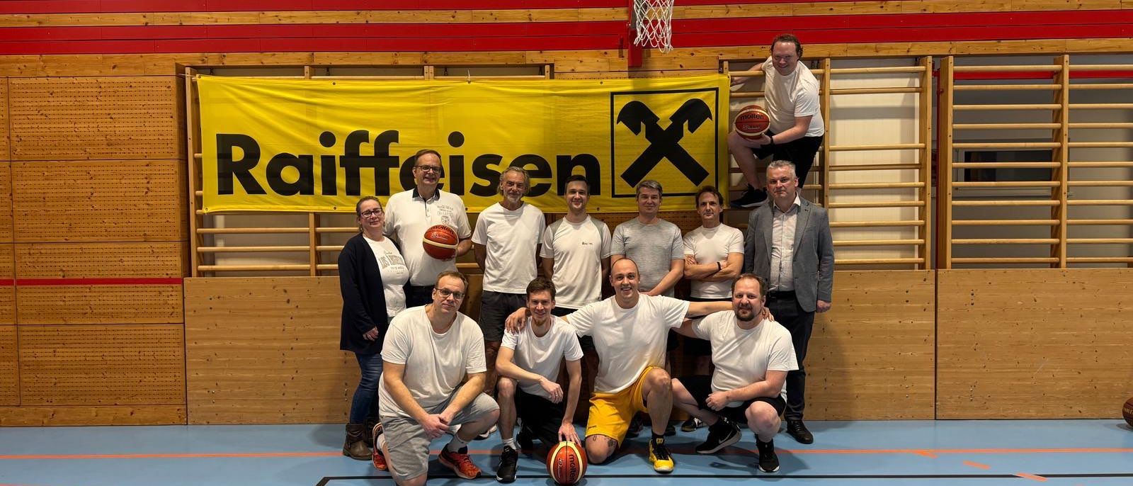 Eine Gruppe von Menschen posiert für ein Foto in einer Turnhalle mit einem Basketballplatz. Ein Banner mit der Aufschrift 'Raiffeisen' hängt über ihnen.