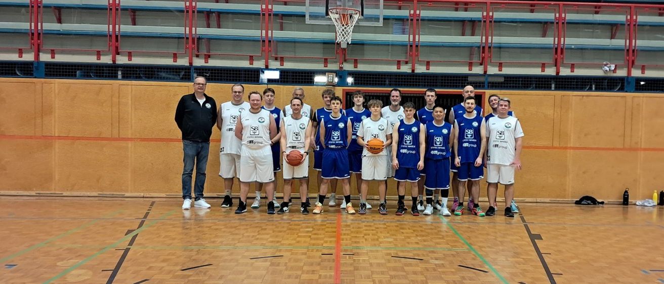 Ein Basketballteam posiert für ein Foto auf einem Hallenspielfeld mit einem Basketballkorb und Tribünen im Hintergrund.