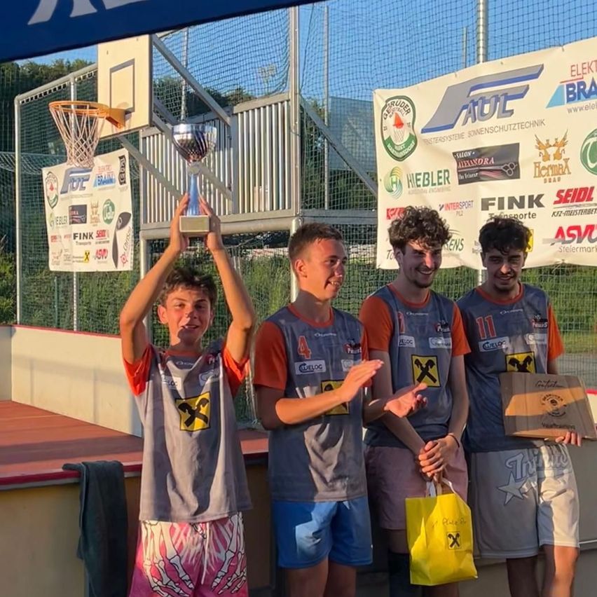 Vier junge Männer stehen lächelnd auf einer Bühne. Einer hält eine Trophäe, ein anderer einen gelben Beutel. Sie tragen Trikots mit Nummern und Buchstaben, und ein Zaun mit Bannern steht hinter ihnen.